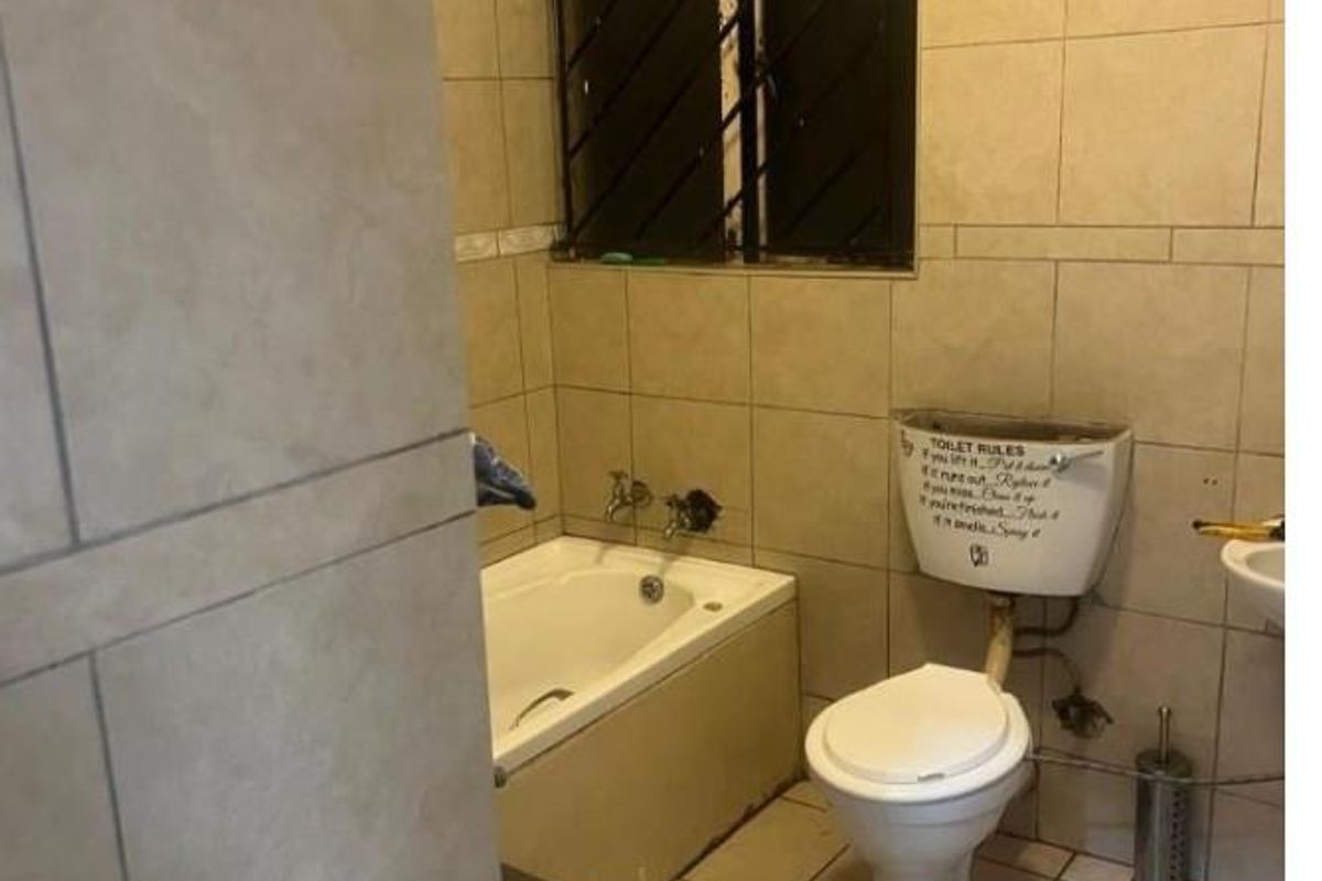 A dated bathroom featuring a built-in bathtub, toilet, and wall-mounted basin with full-height wall tiling and security bars on the window.