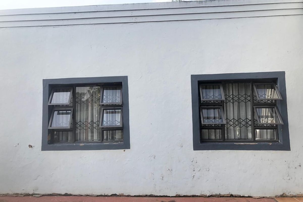 Exterior view of a rear wall featuring two top-hung windows with security bars, showing signs of weathered plaster and paint deterioration.