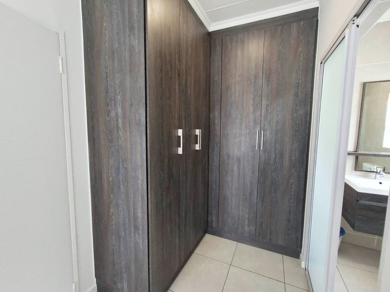 A clean bedroom area featuring dark wood-grain built-in cupboards and neutral floor tiles, with an adjacent view into a modern bathroom.