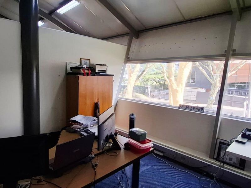A functional office space featuring exposed steel roof beams, large windows providing natural light, and carpeted flooring.
