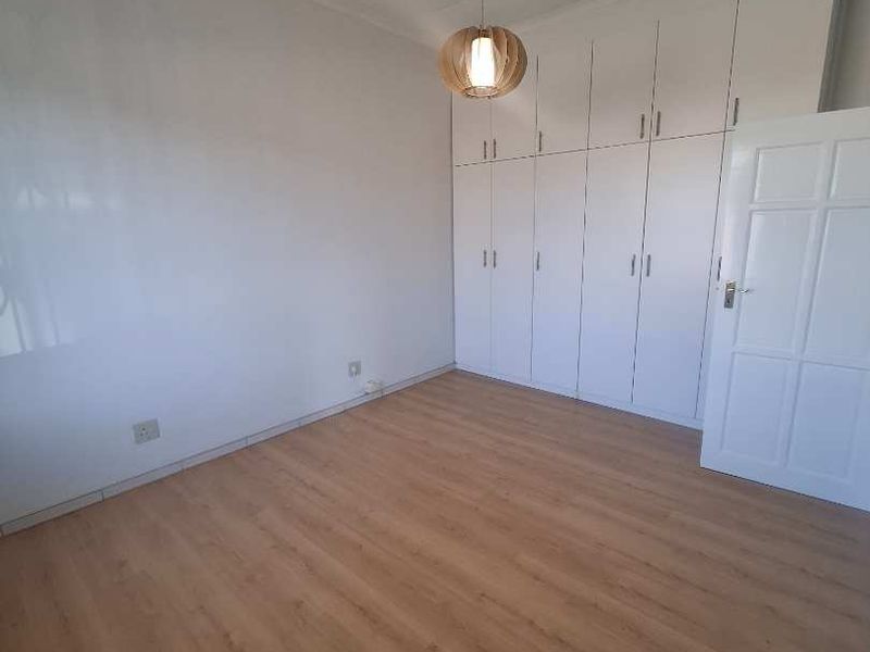 A bright, empty bedroom featuring light wood-look laminate flooring, ample built-in cupboard storage, and a modern pendant light fixture.
