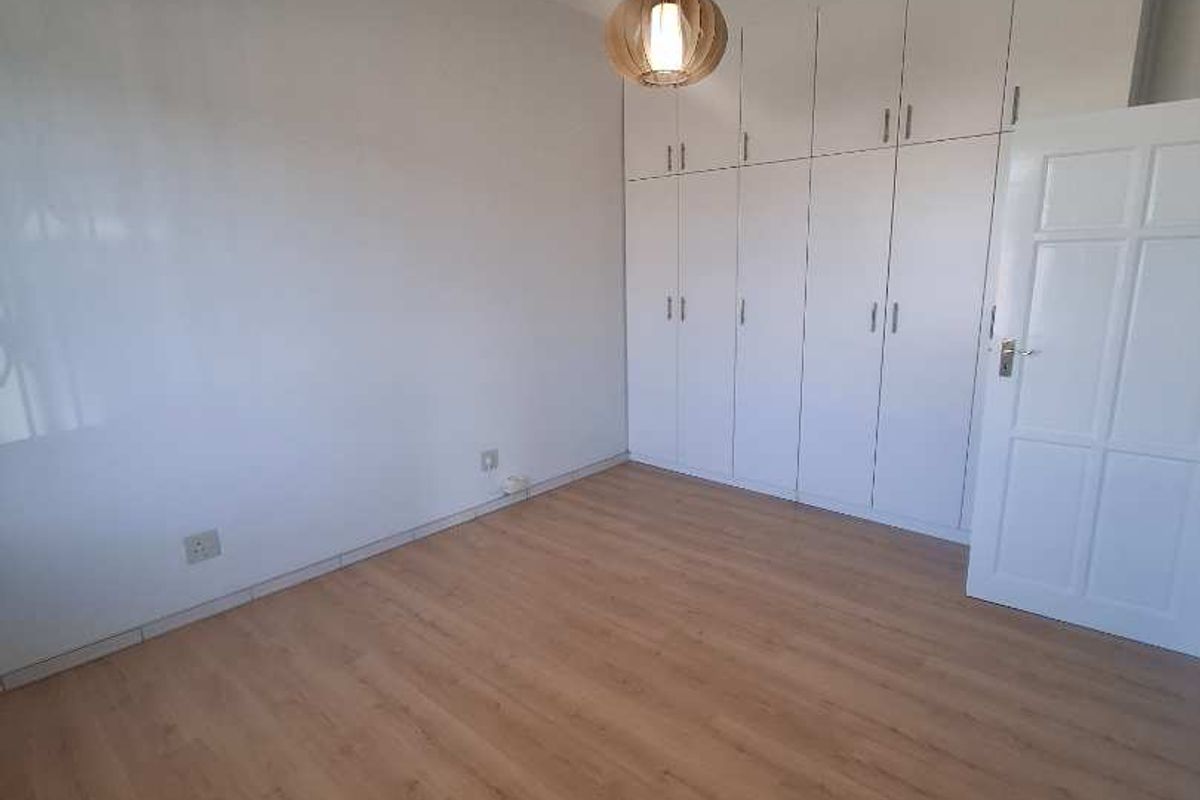 A bright, empty bedroom featuring light wood-look laminate flooring, ample built-in cupboard storage, and a modern pendant light fixture.