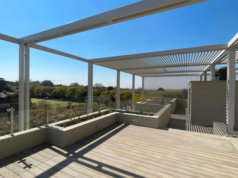 Spacious rooftop balcony featuring timber decking, a modern metal pergola, glass balustrades, and built-in brick planter boxes with panoramic views.