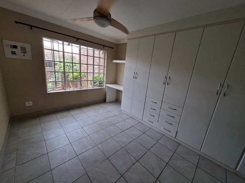 A spacious, empty bedroom featuring tiled flooring, extensive built-in cupboards, a built-in desk, and a ceiling fan.