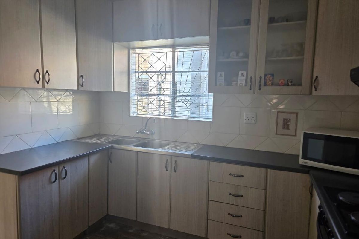 A functional kitchen featuring light-toned cabinetry, laminate countertops, a stainless steel double sink, and tiled backsplash with security bars on the window.