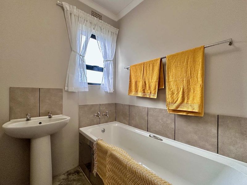 A clean and functional bathroom featuring a pedestal washbasin, a built-in bathtub with tiled splashbacks, and a window for natural light.
