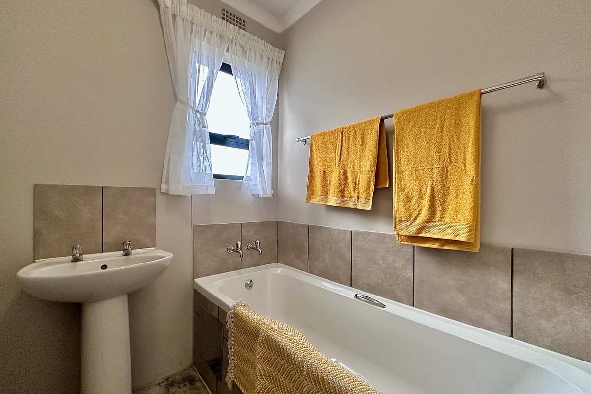A clean and functional bathroom featuring a pedestal washbasin, a built-in bathtub with tiled splashbacks, and a window for natural light.