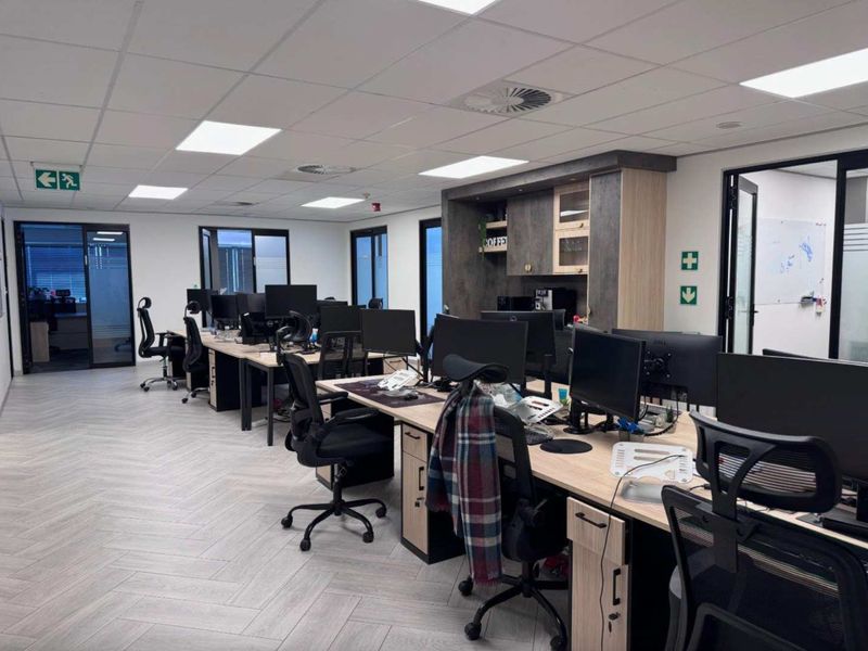 Modern open-plan office space featuring herringbone flooring, suspended ceilings with recessed lighting, and integrated kitchenette cabinetry with glass partition access.