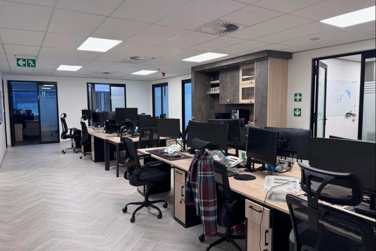 Modern open-plan office space featuring herringbone flooring, suspended ceilings with recessed lighting, and integrated kitchenette cabinetry with glass partition access.
