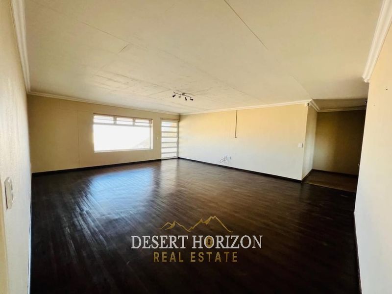 Spacious, empty living area featuring dark wood flooring, ceiling cornicing, and natural light from a large window and sliding door.