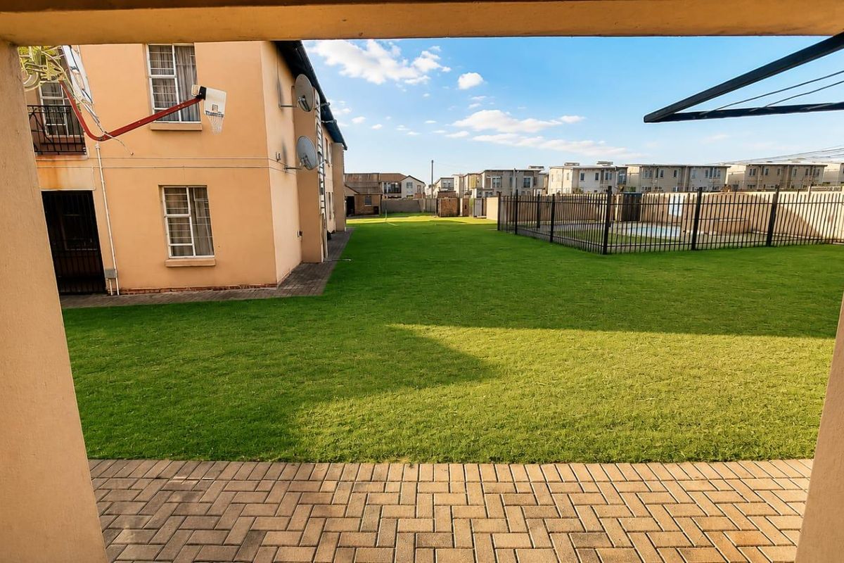 A view of the rear communal garden area featuring a paved patio, well-maintained lawn, and a fenced swimming pool.