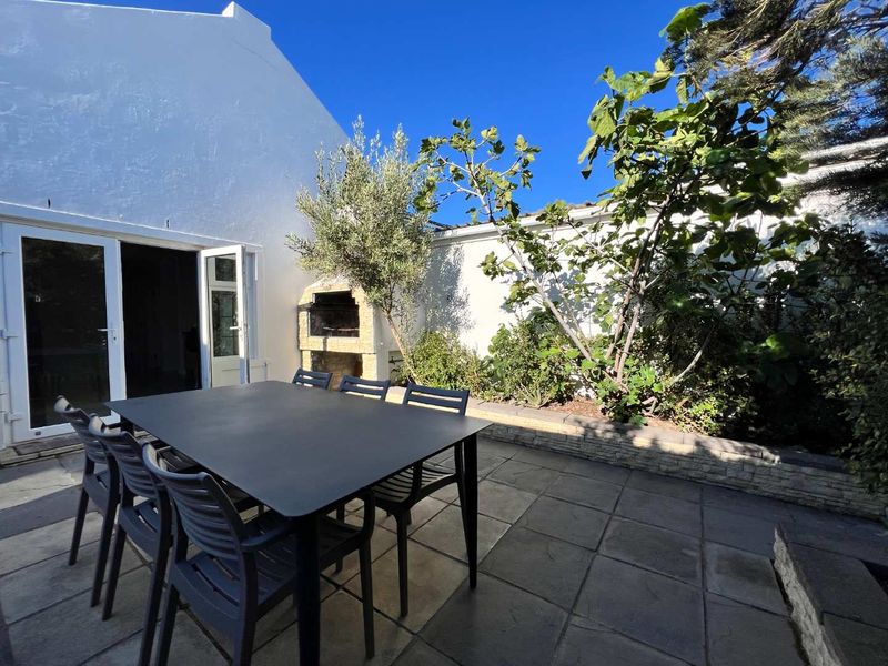 A paved outdoor patio area featuring a built-in braai, established garden greenery, and direct access to the interior through double glass doors.