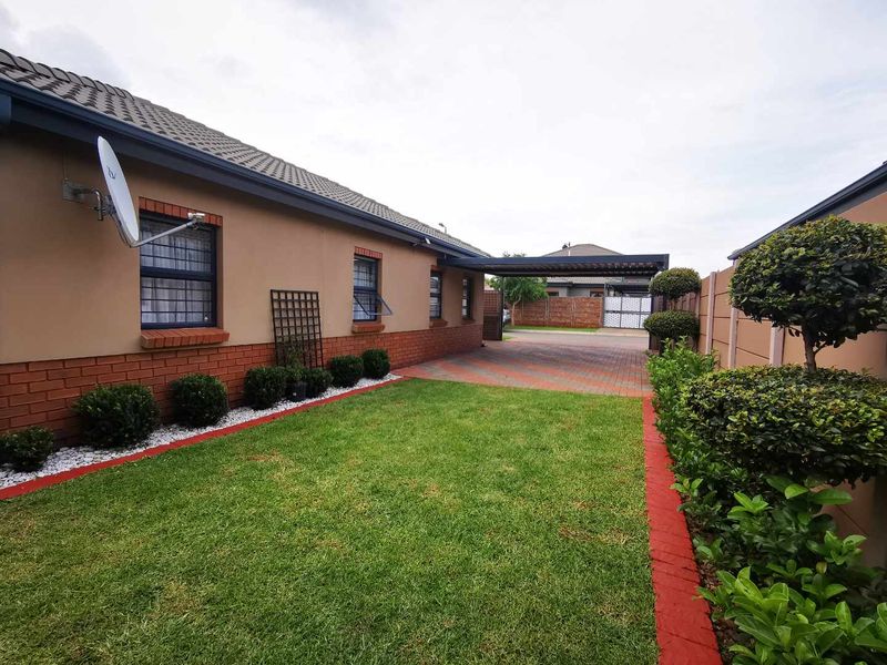 A well-maintained rear exterior featuring a paved driveway, carport, neat lawn, and established garden beds enclosed by a brick boundary wall.