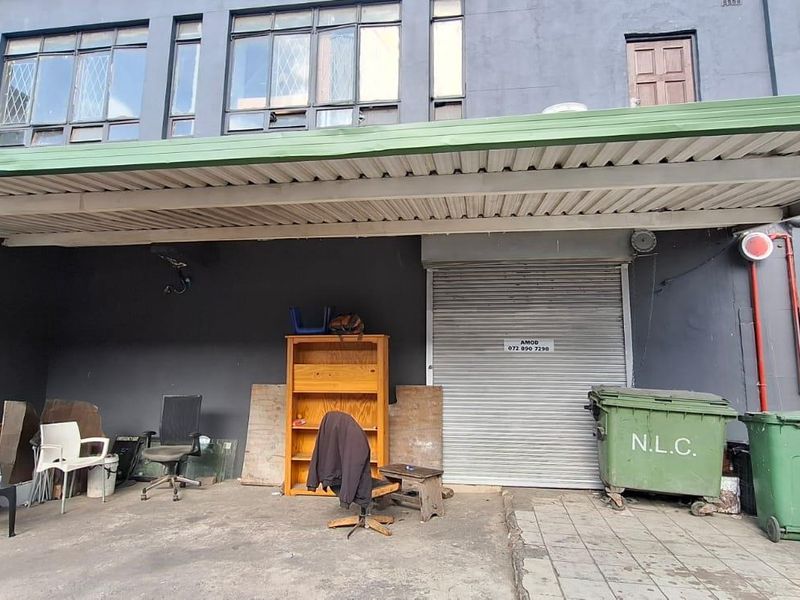 Rear industrial exterior area featuring a metal roller shutter door, corrugated awning, and exposed fire suppression piping against a dark painted facade.