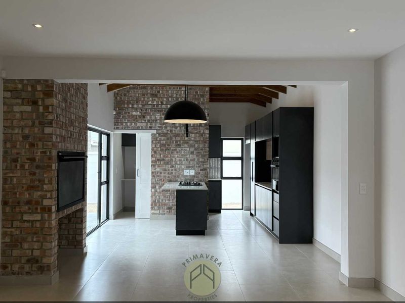 Modern open-plan living and kitchen area featuring exposed brick accents, timber ceiling beams, a central kitchen island with gas hob, and sleek built-in cabinetry.