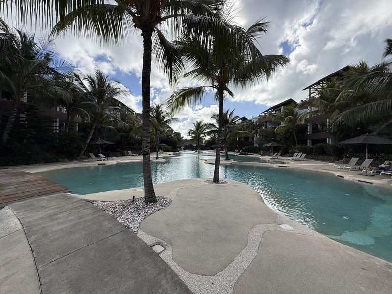A beautifully landscaped communal swimming pool area featuring a large lagoon-style pool, palm trees, and paved walkways within a luxury penthouse complex.