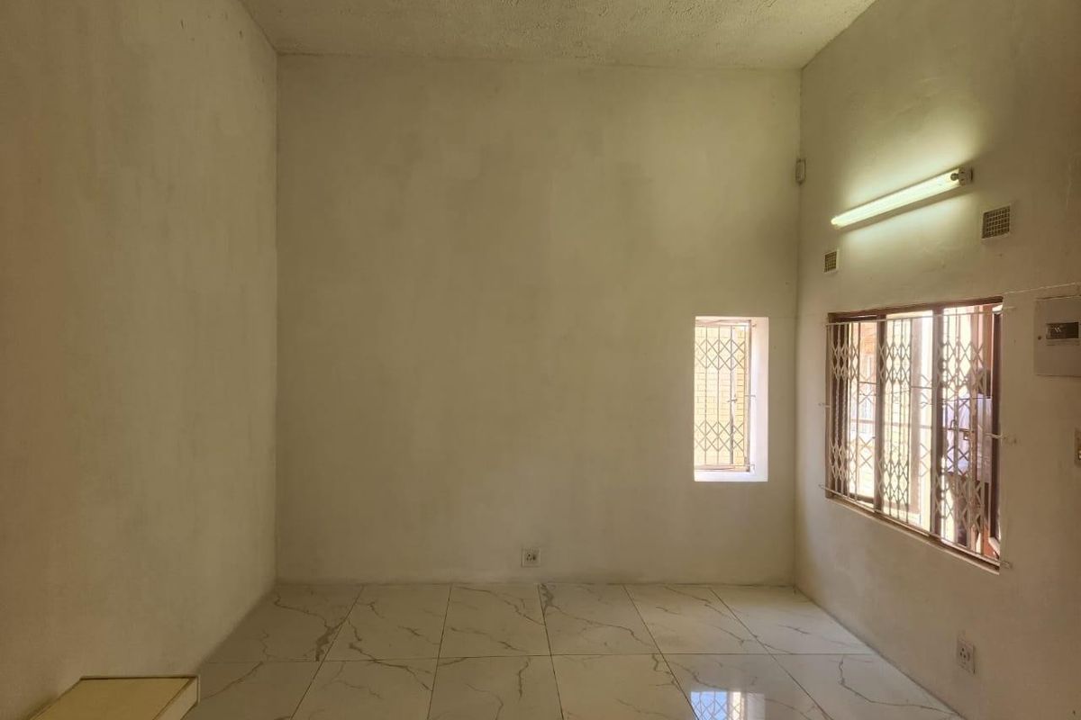 An empty, neutrally finished living area featuring large format floor tiles, security bars on windows, and basic lighting fixtures, ready for personalization.