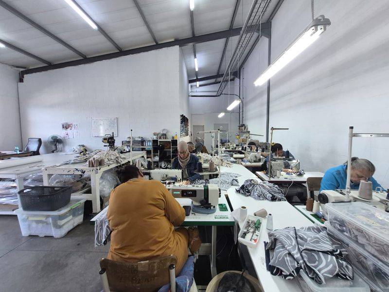 Industrial workshop space featuring high ceilings with exposed steel trusses, concrete flooring, and functional fluorescent lighting, currently utilized as a garment manufacturing facility.