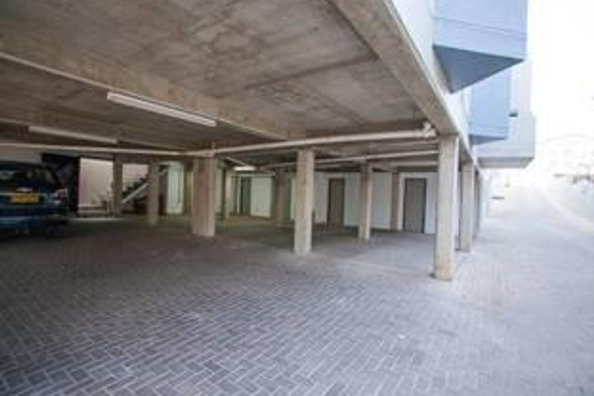 A covered parking area featuring concrete structural columns, paved flooring, and overhead lighting, providing sheltered vehicle access for the office building.