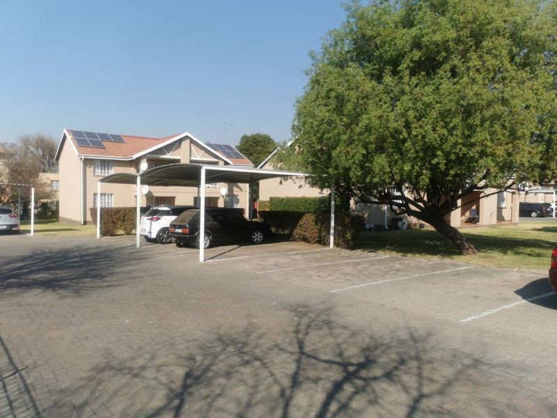 Exterior view of the apartment complex featuring a paved parking area with covered carports, solar panels on the roof, and established garden surroundings.