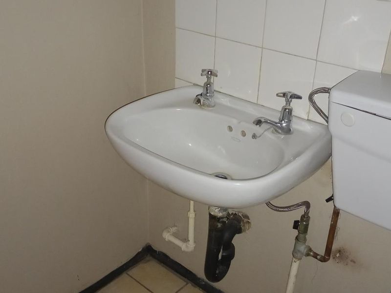 A compact bathroom featuring a wall-mounted basin, tiled splashback, and exposed plumbing fixtures, requiring cosmetic updates to improve its overall condition.