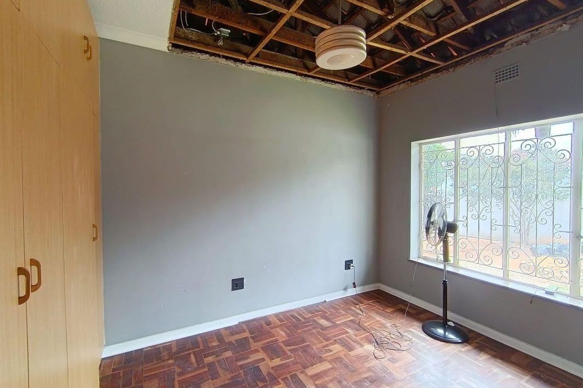 A bedroom featuring parquet flooring, built-in cupboards, and a window with security bars, currently requiring ceiling repairs.
