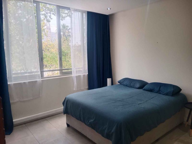 A bright bedroom featuring large windows with natural light, recessed downlights, and neutral tiled flooring.