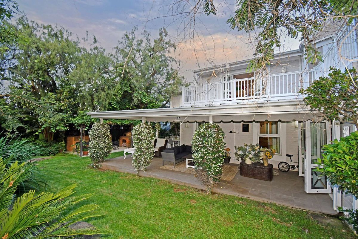 A spacious rear garden featuring a covered patio area, a built-in brick braai, and an upper-level balcony overlooking the lawn.