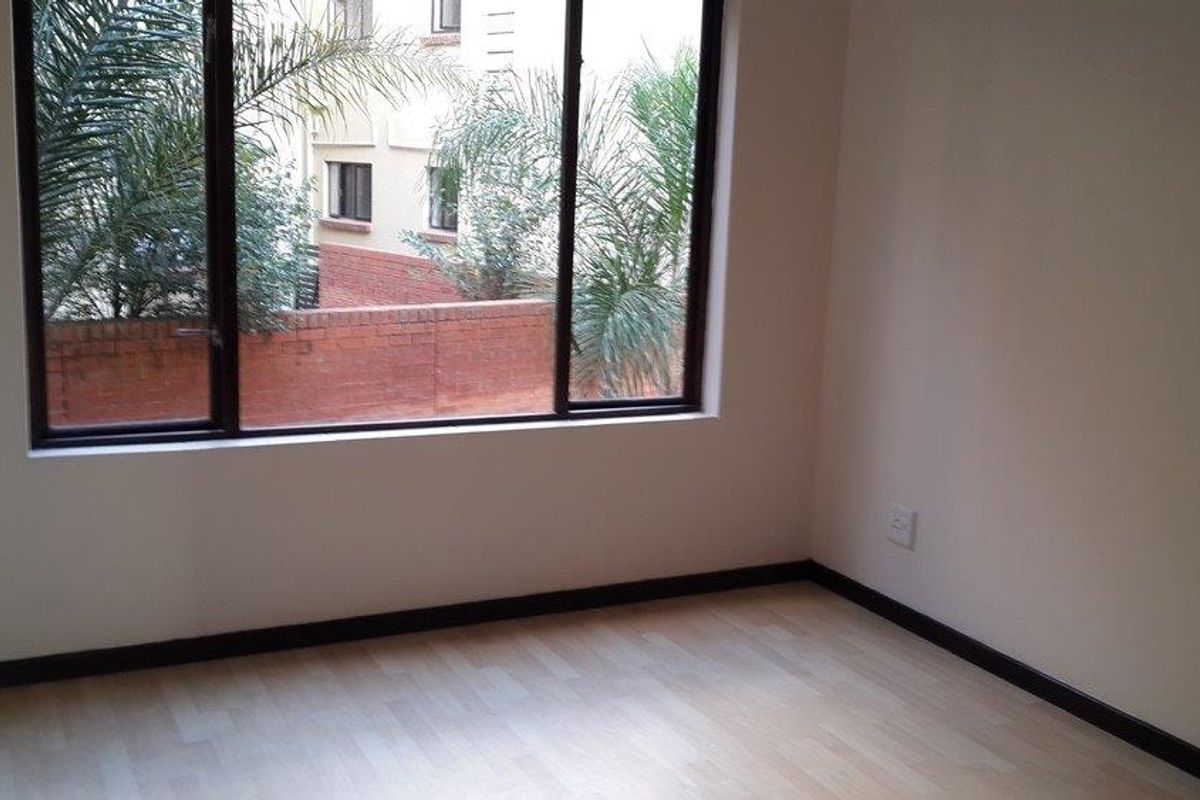 A bright, empty bedroom featuring light-toned laminate flooring, dark skirting boards, and large windows overlooking a brick-walled exterior area.