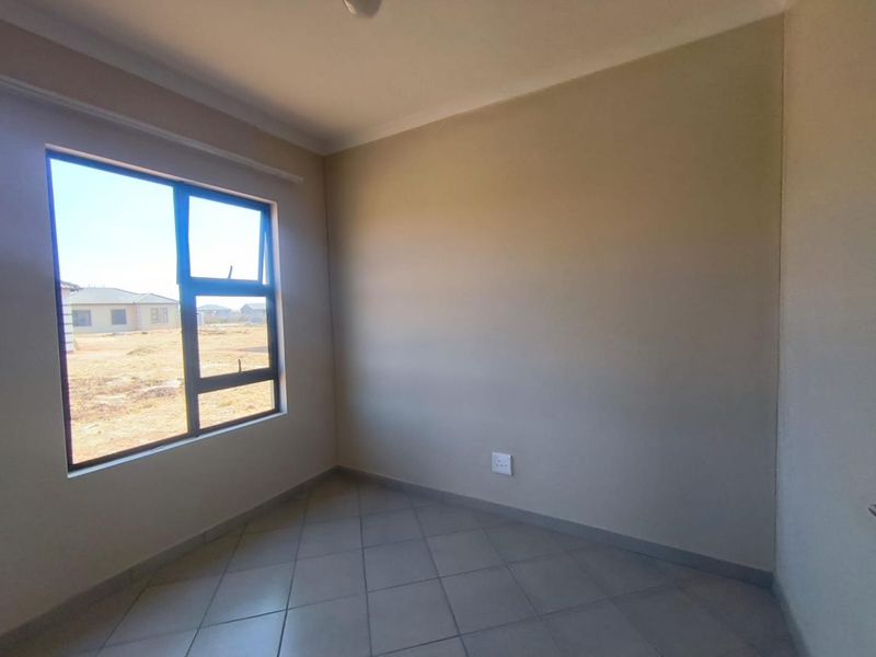 A clean, empty bedroom featuring neutral wall paint, tiled flooring, and a large aluminium-framed window providing natural light.