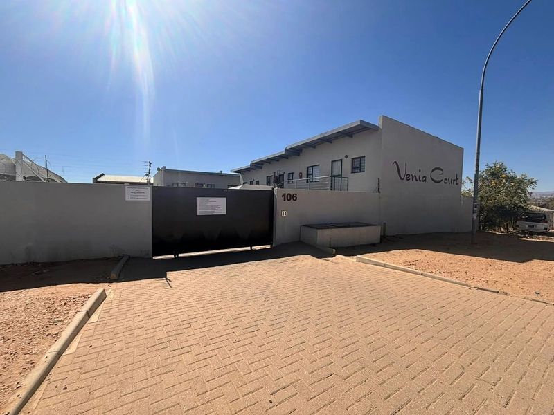 Front exterior view of Venia Court townhouse complex featuring a paved driveway, secure perimeter wall with electric fencing, and a large sliding entrance gate.