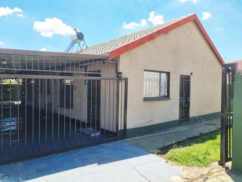 Front exterior view of the house featuring a paved driveway, metal carport, security gates, and a roof-mounted solar water heater.