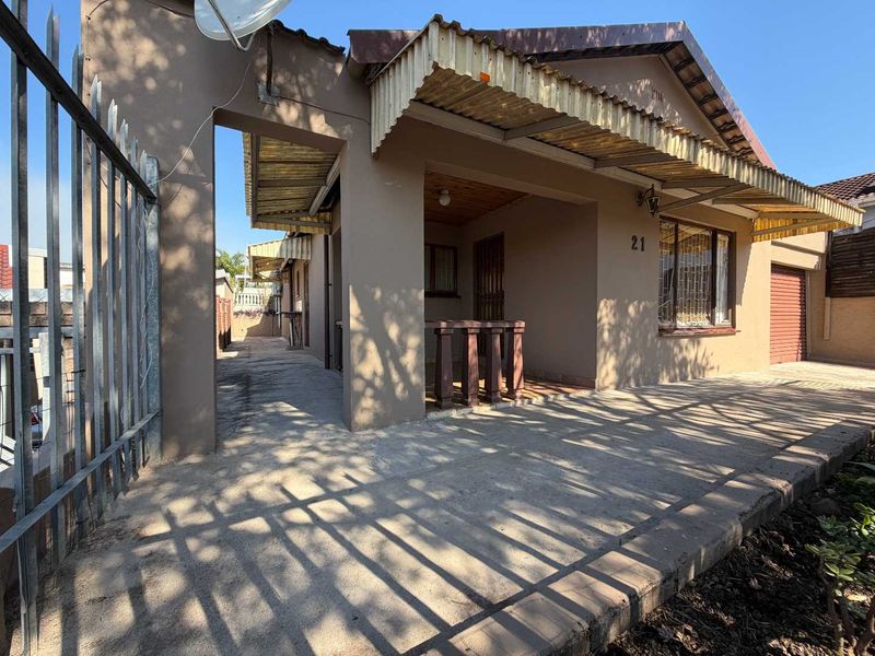 Front elevation of a single-story house featuring a concrete driveway, security fencing, and a corrugated metal awning over the entrance area.