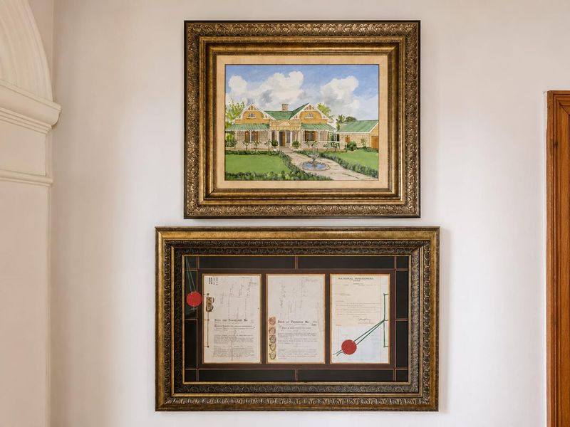 A wall display featuring a framed painting of a house and a framed collection of historical property deeds.