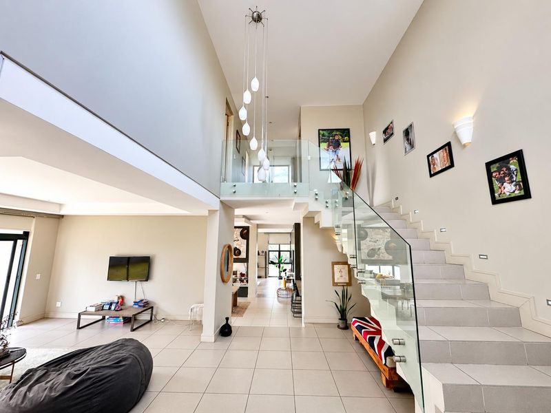Spacious open-plan living area featuring a striking double-volume ceiling, a modern staircase with glass balustrades, and large format floor tiles.