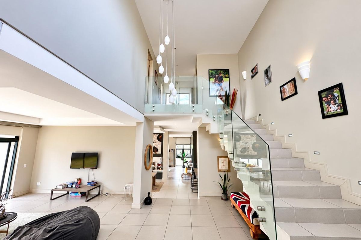 Spacious open-plan living area featuring a striking double-volume ceiling, a modern staircase with glass balustrades, and large format floor tiles.