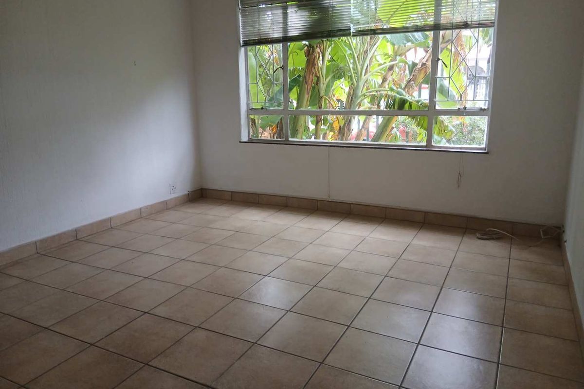 A vacant bedroom featuring neutral ceramic floor tiles, a large window with fitted blinds, and a view of lush greenery.