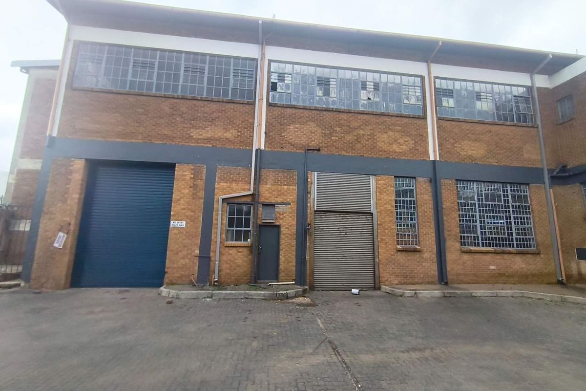 Industrial rear facade featuring brick construction, multiple roller shutter doors, and steel-framed windows, set within a paved yard area.