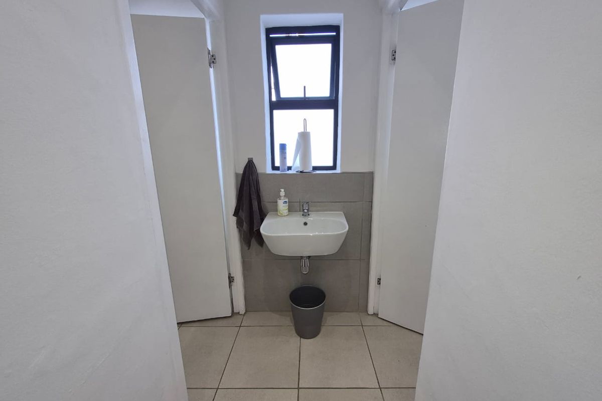 A clean and functional office bathroom featuring a wall-mounted basin, tiled splashback, and neutral floor tiles with natural light from a casement window.