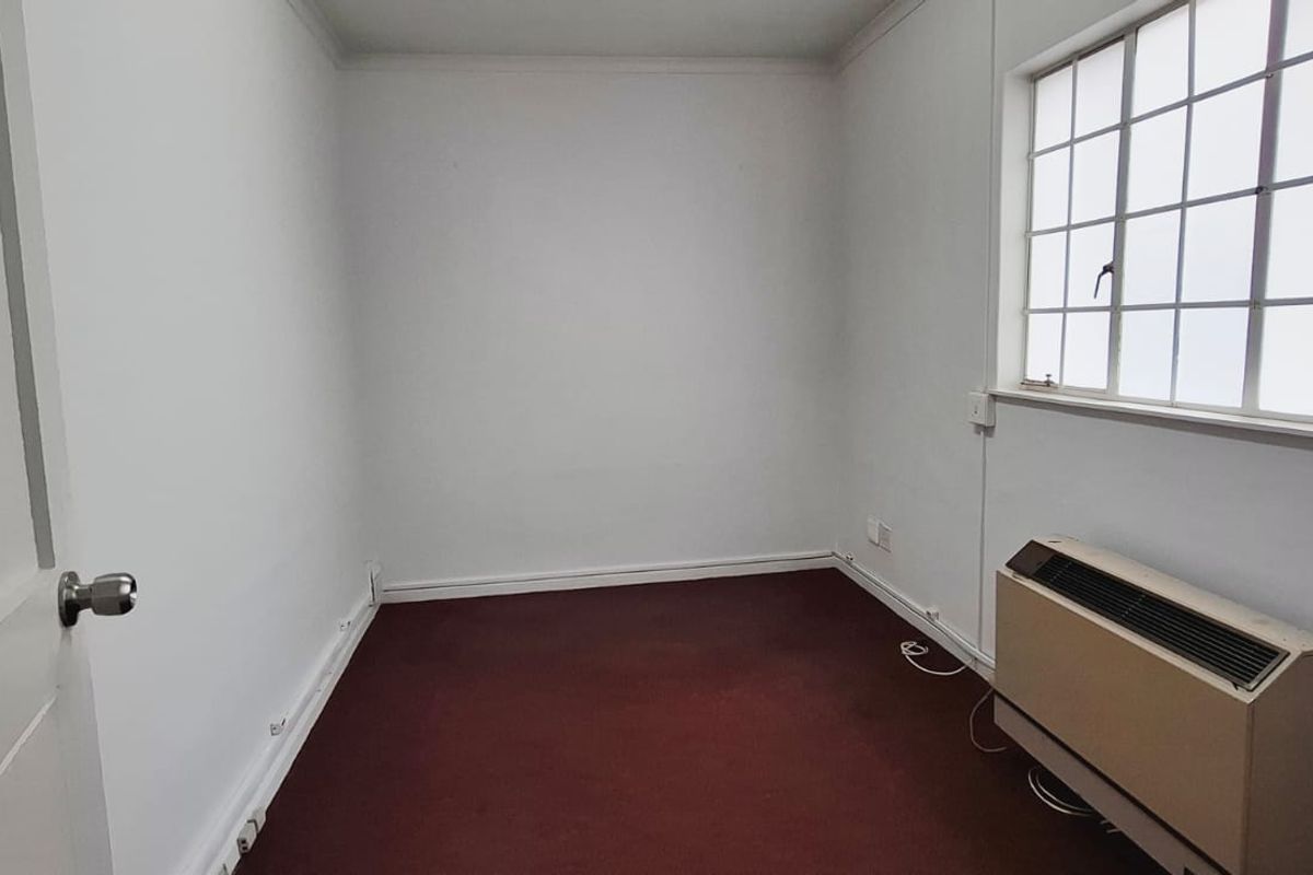 A vacant office space featuring white walls, carpet flooring, a large grid window, and a wall-mounted HVAC unit.