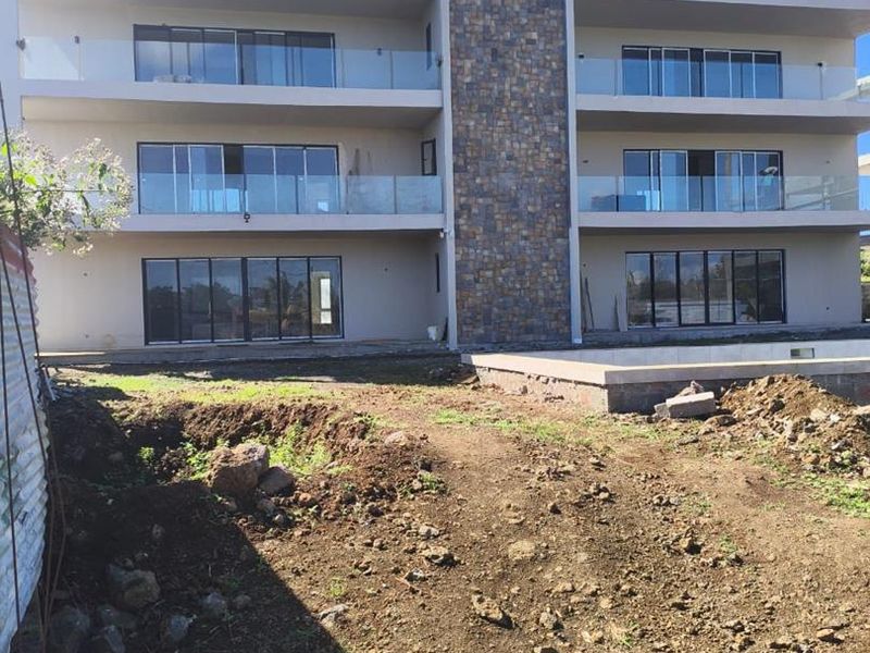 Rear view of a modern apartment building featuring glass balustrades, stone cladding, and ground-level access to a swimming pool area with unfinished landscaping.