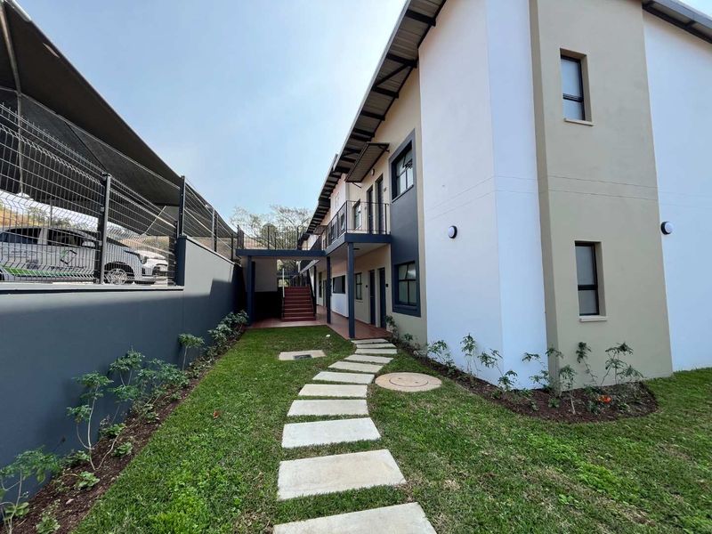 A well-maintained rear exterior view of the apartment complex featuring a landscaped garden, paved walkway, and access to upper-level balconies via a metal staircase.