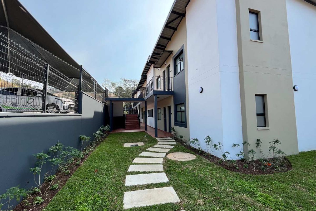 A well-maintained rear exterior view of the apartment complex featuring a landscaped garden, paved walkway, and access to upper-level balconies via a metal staircase.