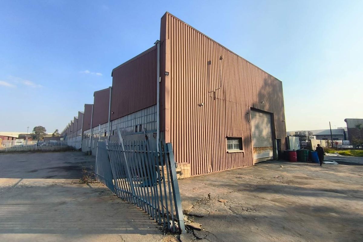 Exterior view of an industrial warehouse featuring corrugated metal cladding, a large roller shutter door, and surrounding concrete yard space with perimeter fencing.