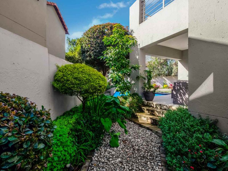 A landscaped rear garden featuring a stone pathway leading to a covered patio area with a swimming pool and balcony above.