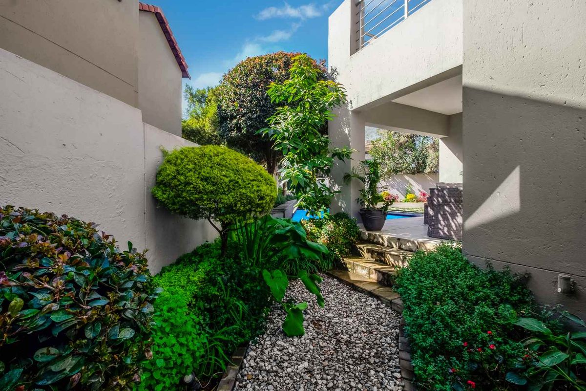 A landscaped rear garden featuring a stone pathway leading to a covered patio area with a swimming pool and balcony above.