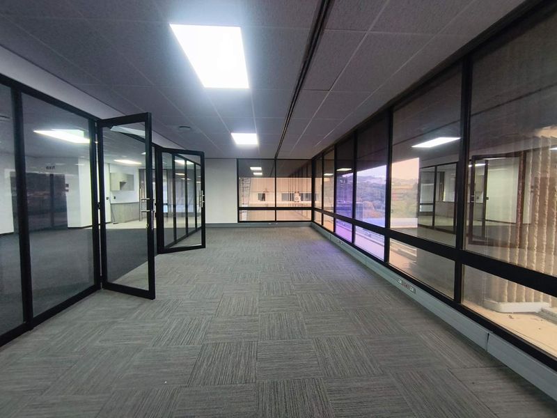 A spacious, vacant office area featuring glass partitions, carpet tiles, and large windows providing natural light and views.