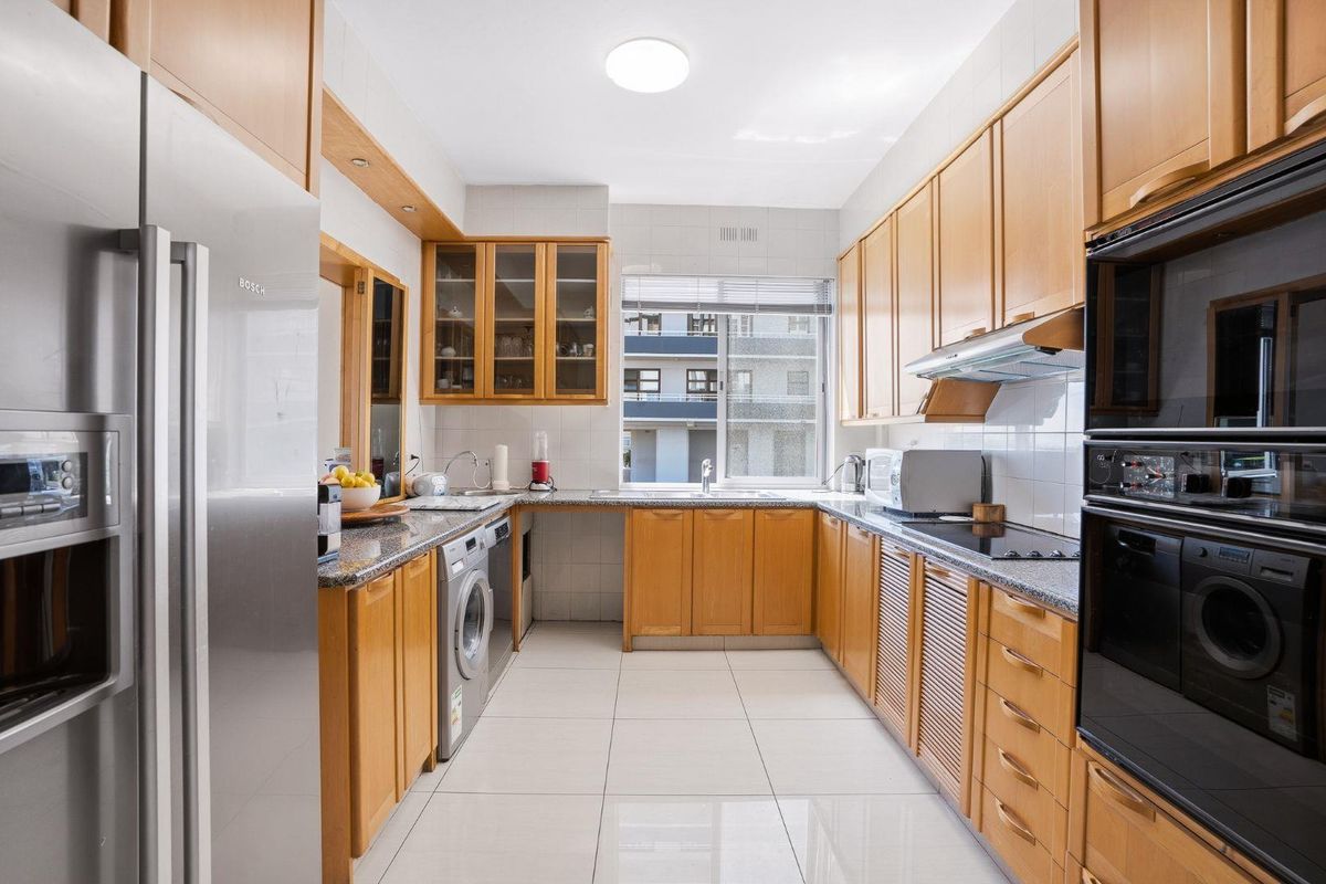 A functional U-shaped kitchen featuring warm-toned wooden cabinetry, granite countertops, integrated appliances, and neutral tiled flooring with ample natural light.