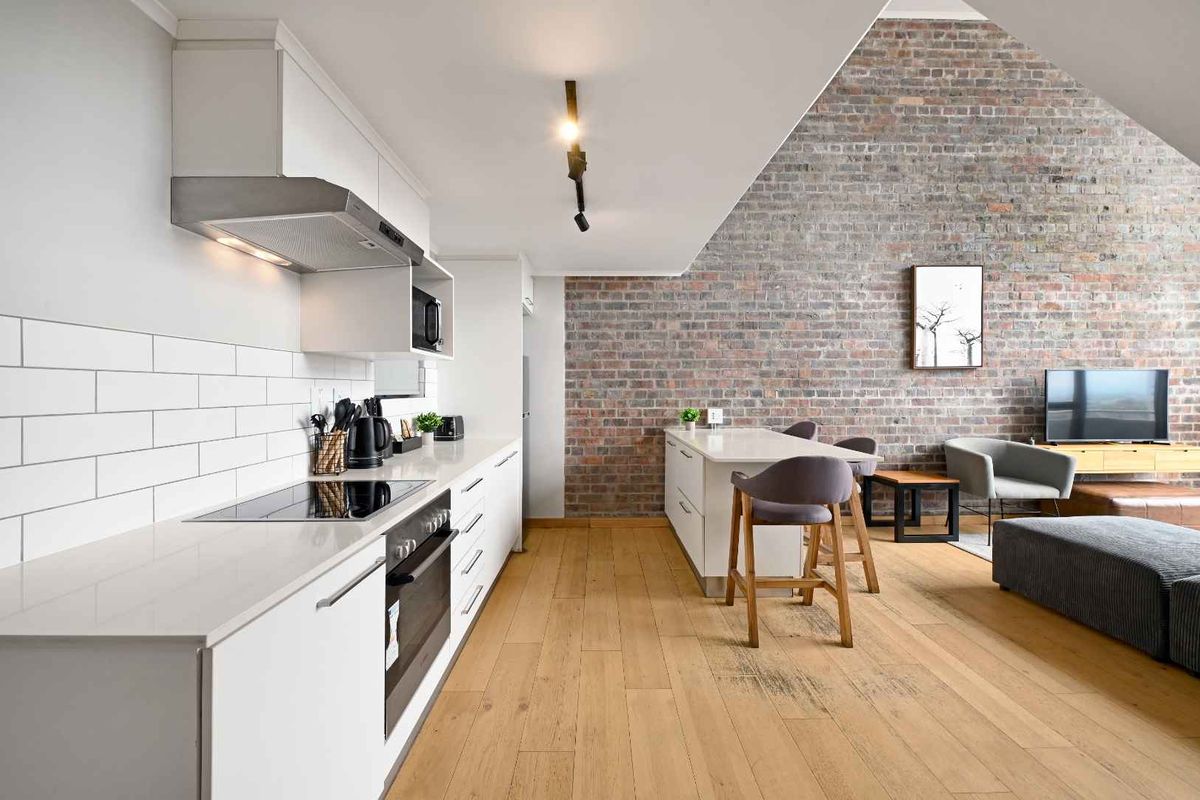 Modern open-plan living area featuring a sleek kitchen with built-in appliances, subway tile backsplash, and a striking exposed brick feature wall.