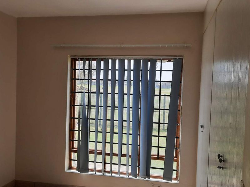 A vacant bedroom featuring built-in cupboards, security bars on the window, and vertical blinds in need of repair.